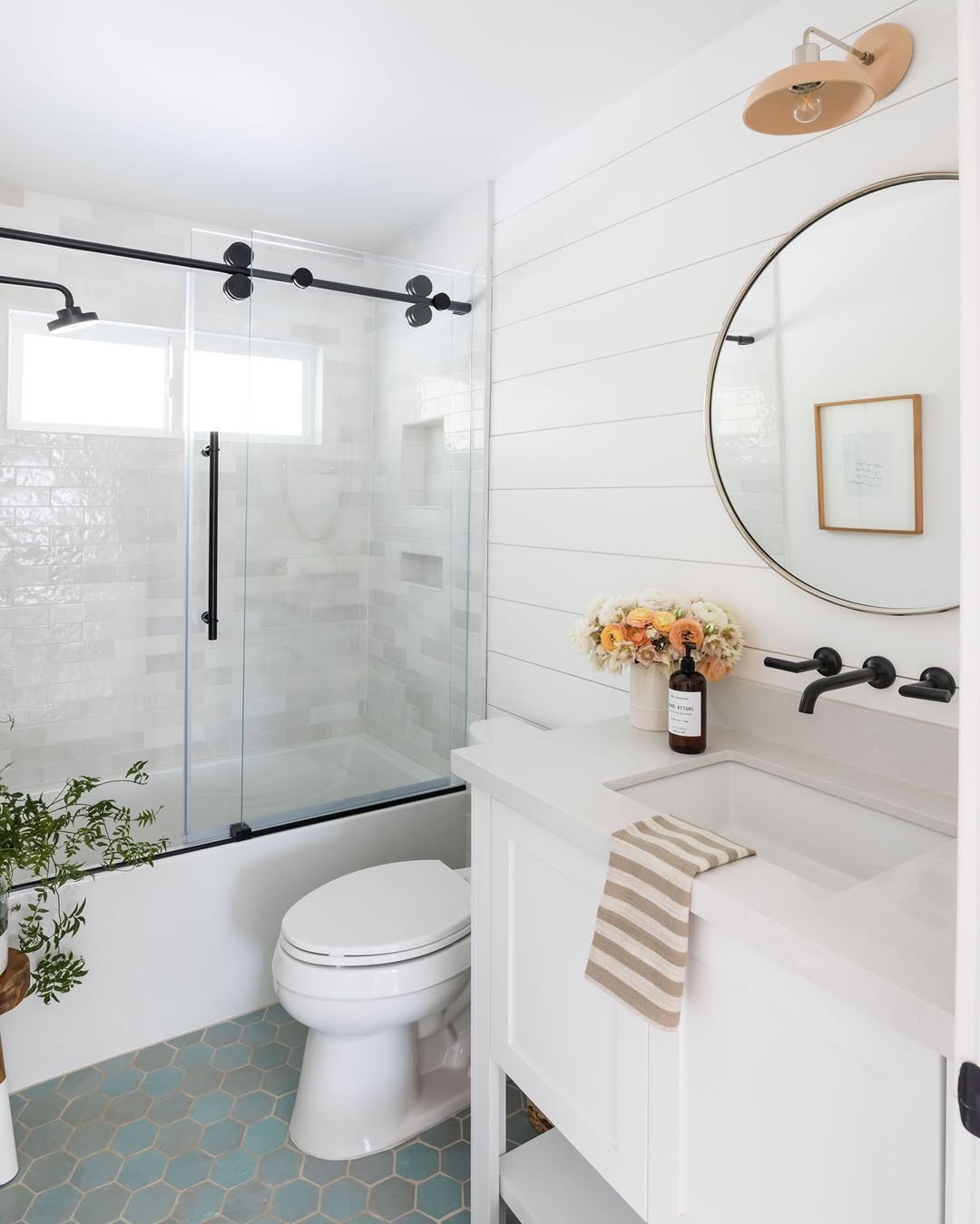 Modern shiplap wall bathroom with a glass shower enclosure, white vanity, round mirror, wall-mounted faucet, striped towel, floral arrangement, and hexagonal tile floor.