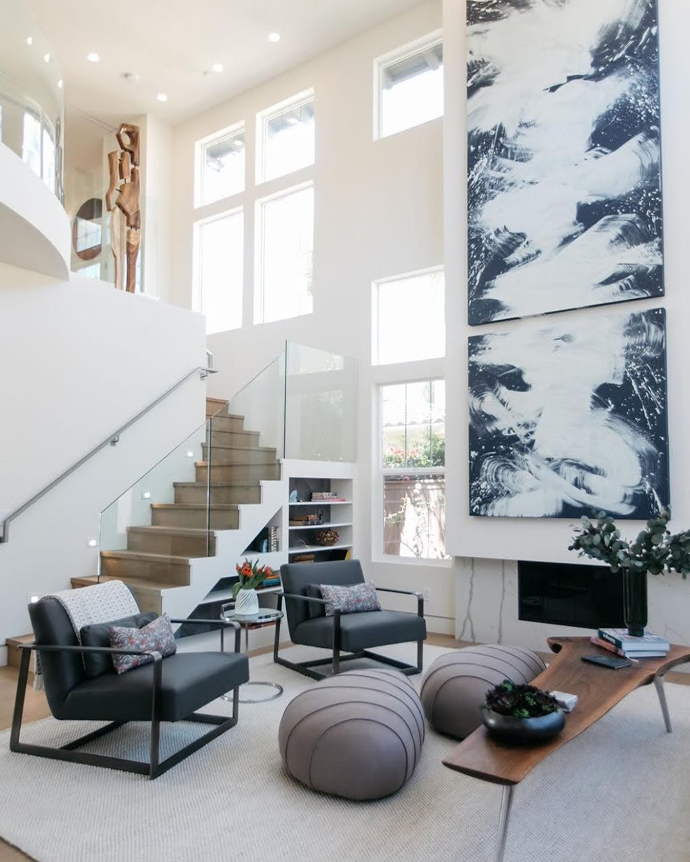 Modern living room with large windows, abstract art above the fireplace, gray chairs, round ottomans, a wooden coffee table, and a staircase with glass railing—perfect for those seeking high ceiling living room ideas.