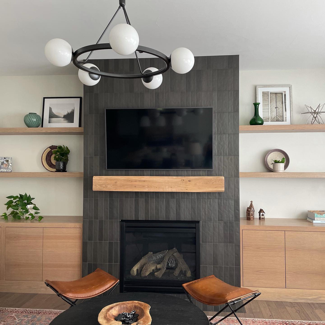 Modern living room with striking fireplace tiles on a black tiled fireplace, mounted TV, wooden mantel, built-in shelves with decor, and two leather chairs around a round coffee table.