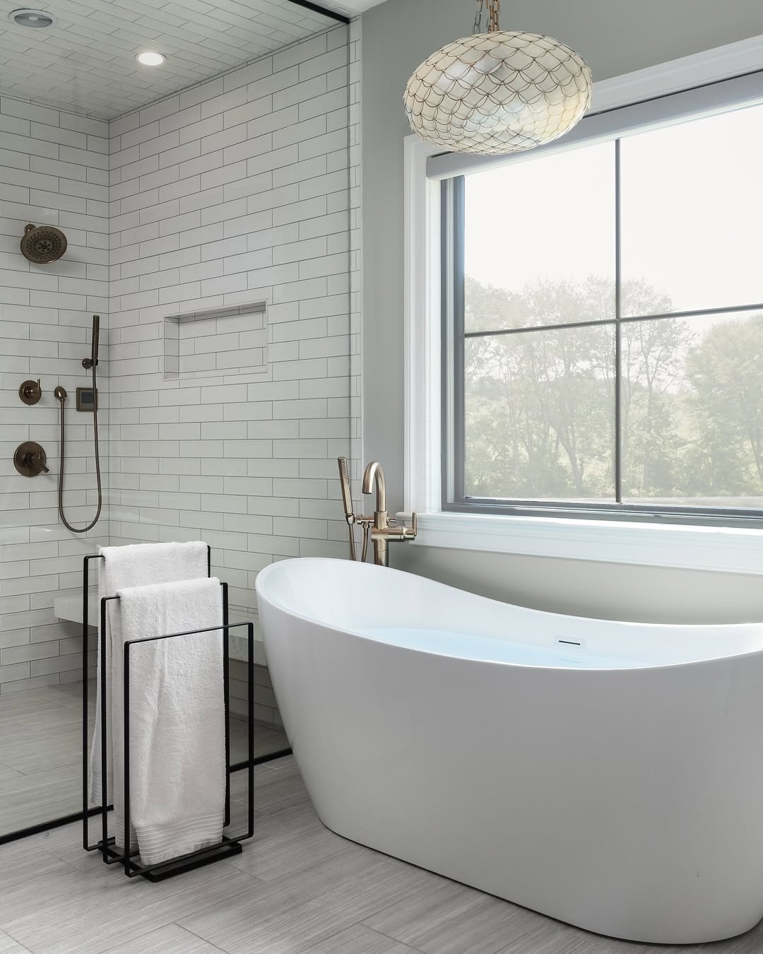 Modern bathroom with a freestanding white bathroom tub, floor-mounted faucet, towel rack, tiled shower with glass wall, large window, and pendant light. Perfect inspiration for those seeking stylish bathroom tub ideas.