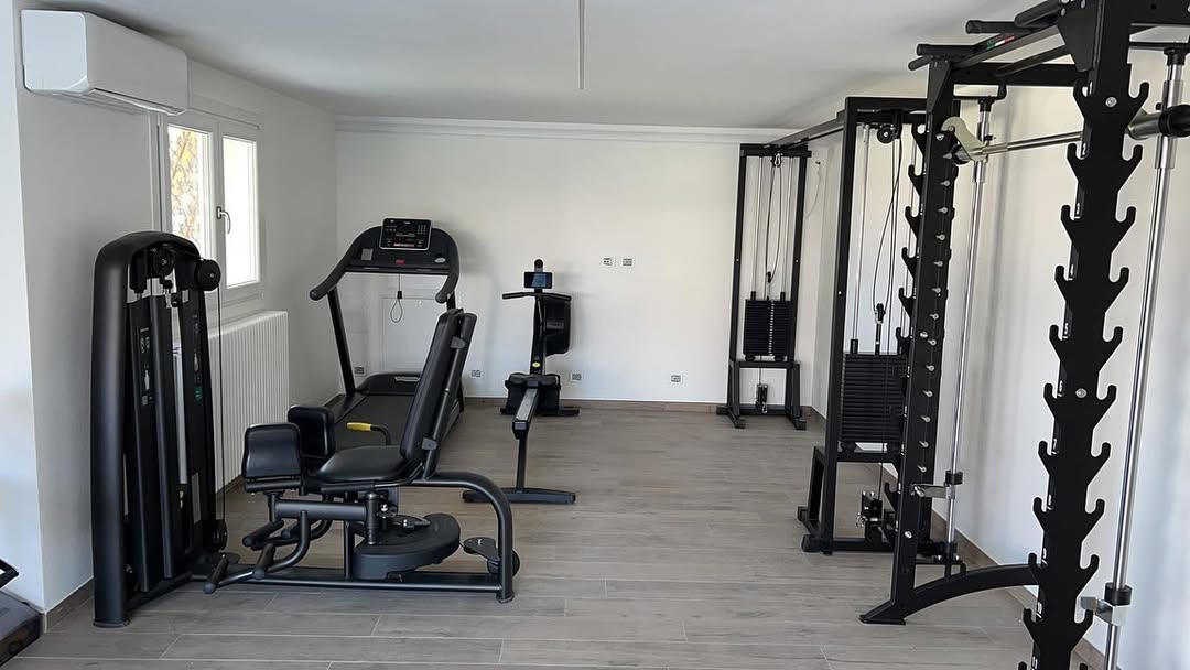 A mini gym at home featuring various exercise machines, including a leg press, treadmill, cable machine, and weight rack, all set up in a bright room with wood flooring.