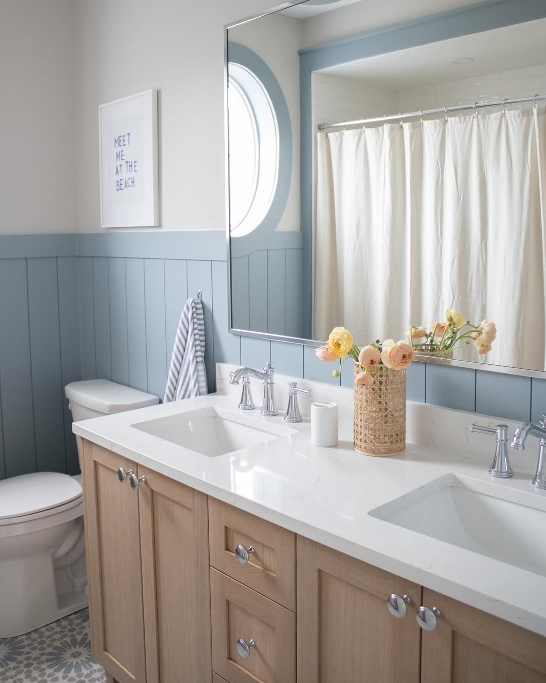 A clean shiplap wall bathroom features a wood vanity with double sinks, a large mirror, a toilet, a striped towel, a shower with a white curtain, and yellow flowers in a wicker vase.