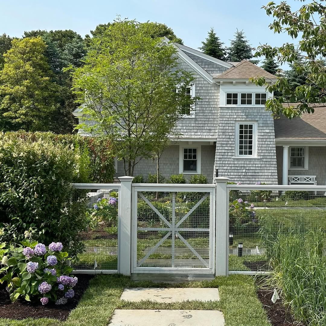 A white farmhouse gate with mesh fencing opens to a stone path leading to a gray shingle house, surrounded by green trees and blooming hydrangeas.