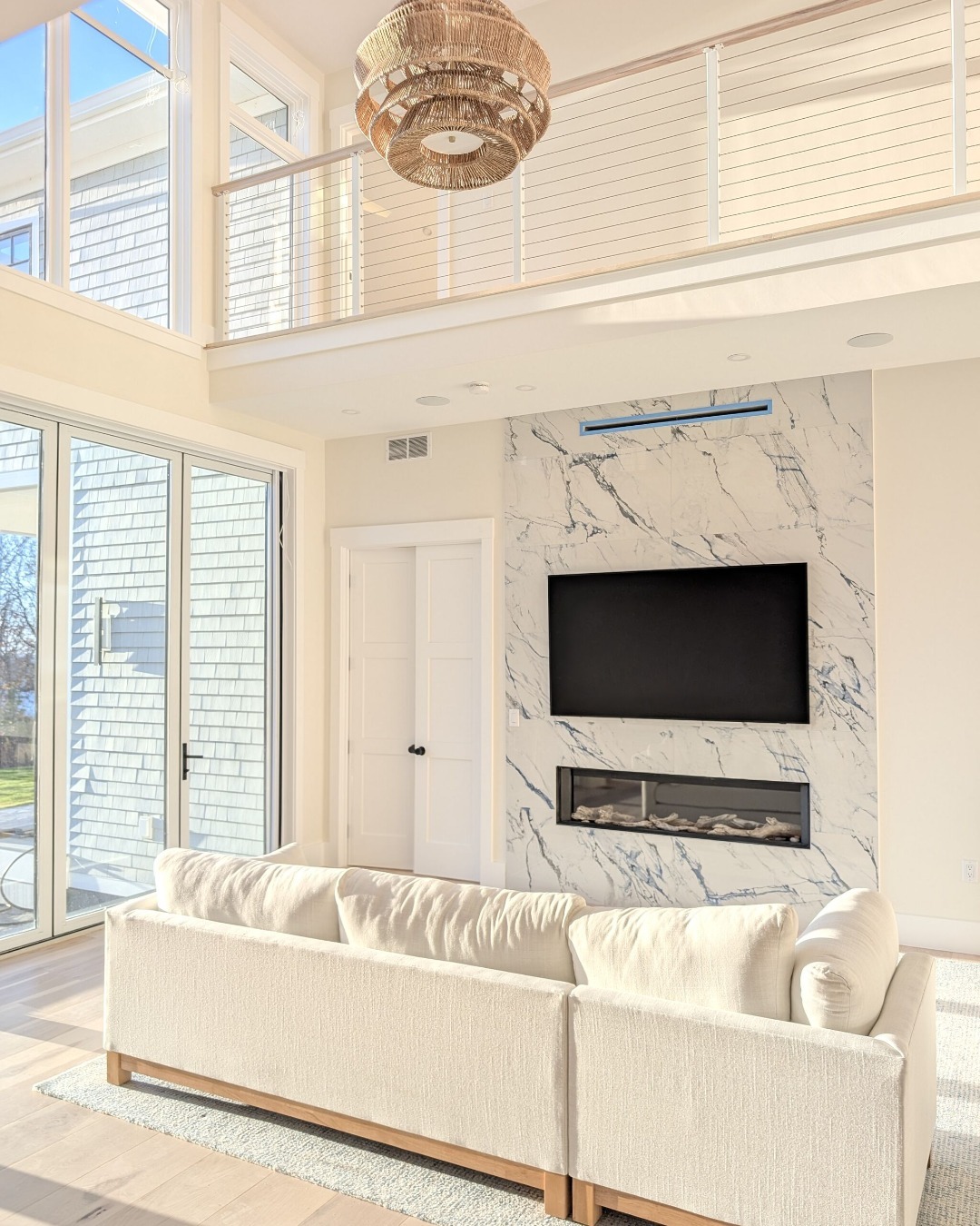 A modern high ceiling living room with a white sofa, marble accent wall featuring a mounted TV and fireplace, large windows, and a balcony with metal railings above.