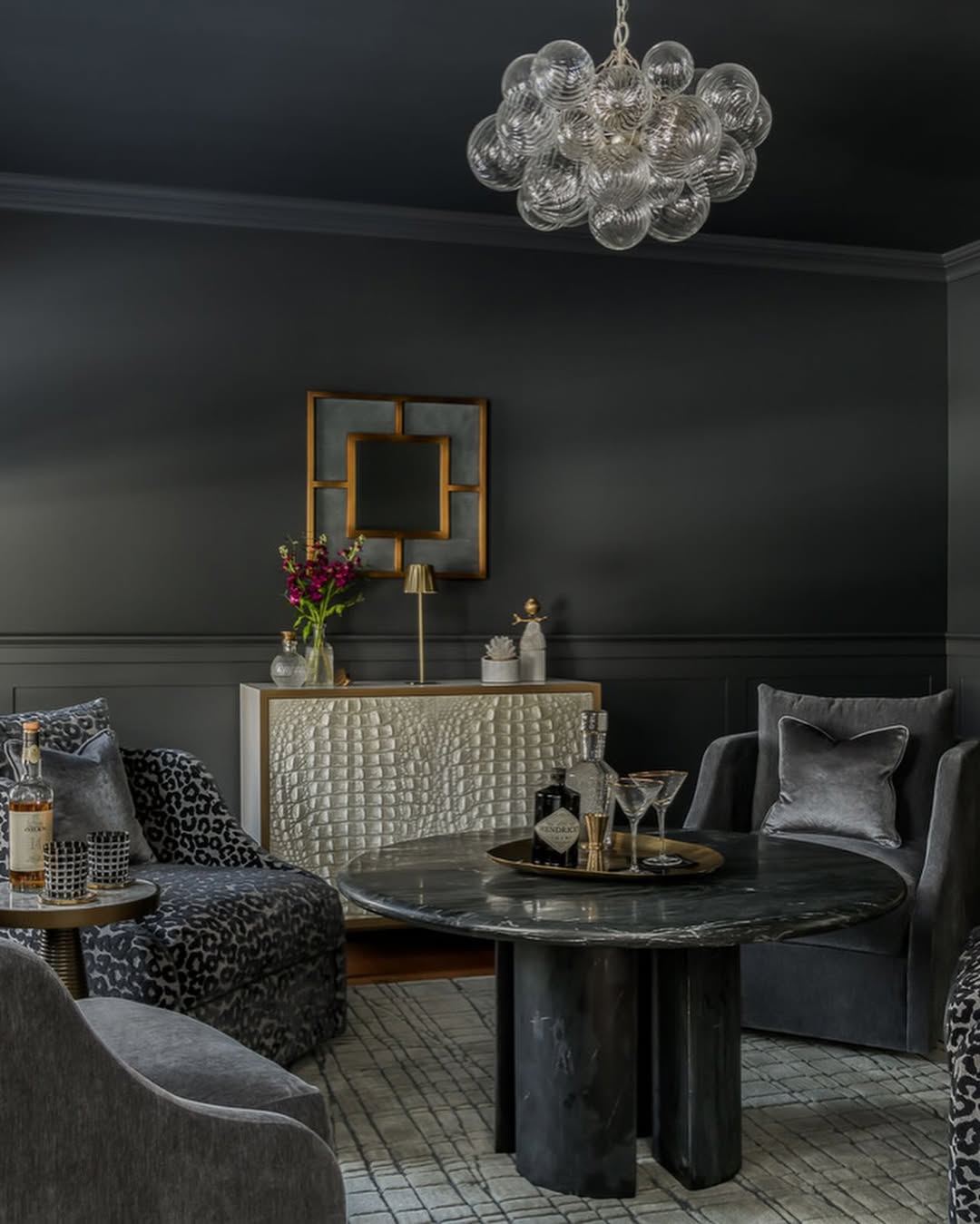 A modern black living room with dark walls, velvet armchairs, a round black marble table, a textured sideboard, and a glass bubble chandelier.