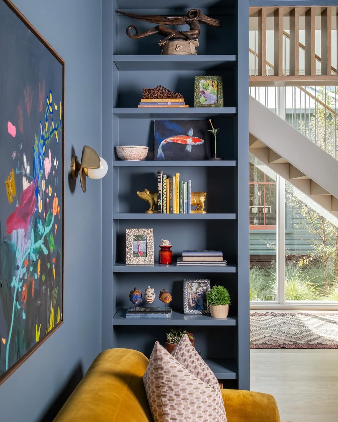 A built-in blue bookshelf showcases creative bookshelf decor, displaying books, framed photos, small art objects, and plants next to a yellow sofa in a modern living room. Stairs and windows are visible in the background.
