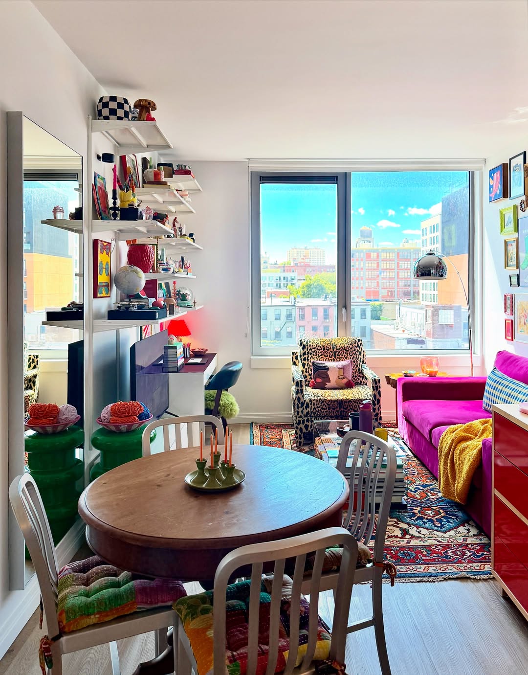 Colorful apartment interior with a round dining table, metal chairs, shelves with decor, a pink sofa, and large window overlooking city buildings—perfect for inspiring small space decorating and tiny apartment ideas.