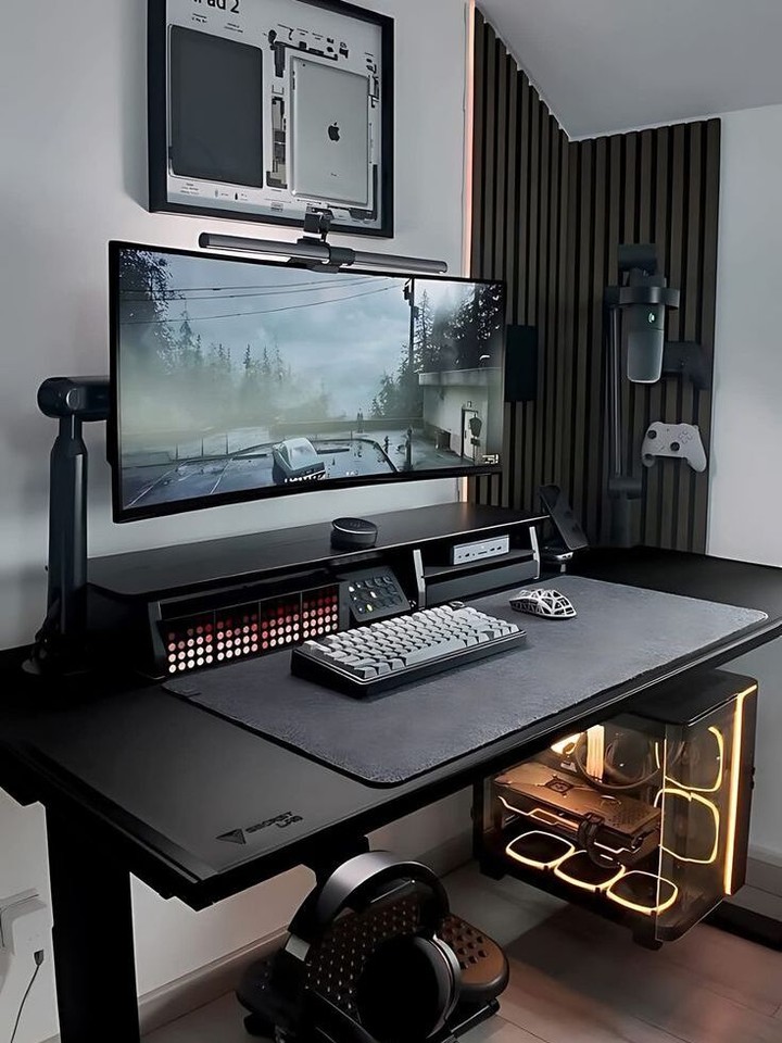 A modern gaming room setup features a large monitor, mechanical keyboard, mouse, and headset on a sleek black desk, with a transparent PC case and mounted accessories—perfect for those seeking the latest gaming room ideas.