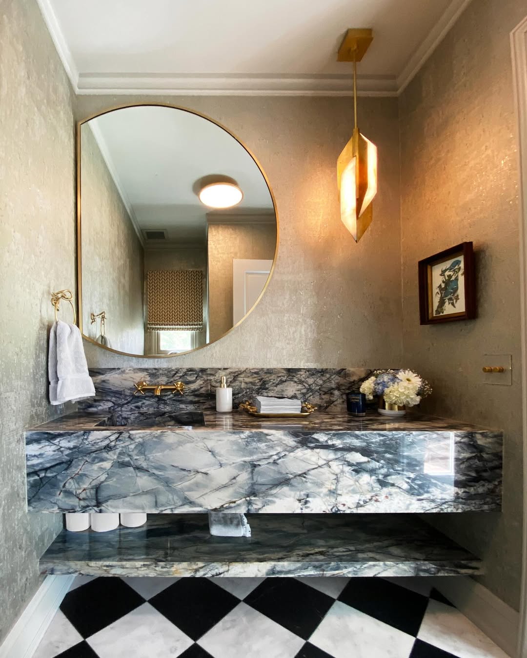 Bathroom with a round mirror, marble sink and shelf, gold fixtures, geometric pendant light, towel, flowers, and a framed picture on textured walls with a black-and-white checkered floor—perfect for stylish powder rooms.