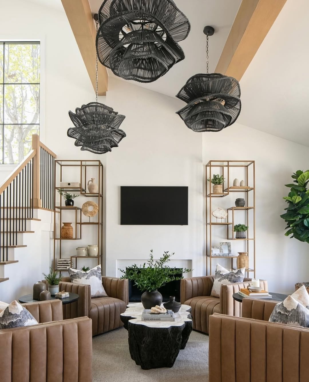 Modern high ceiling living room with tan tufted chairs, black pendant lights, a black coffee table, wall-mounted TV, open shelving with decor, a large plant, and a staircase with wooden railing for that bigger home feel.