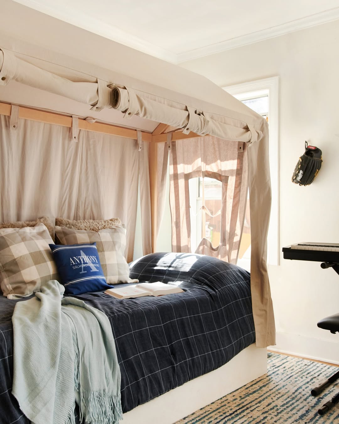 A cozy boys bedroom idea featuring a four-poster bed draped in light fabric, plaid pillows, a blanket, smart storage solutions, a keyboard, a wall-mounted baseball glove, and natural light streaming through the window.