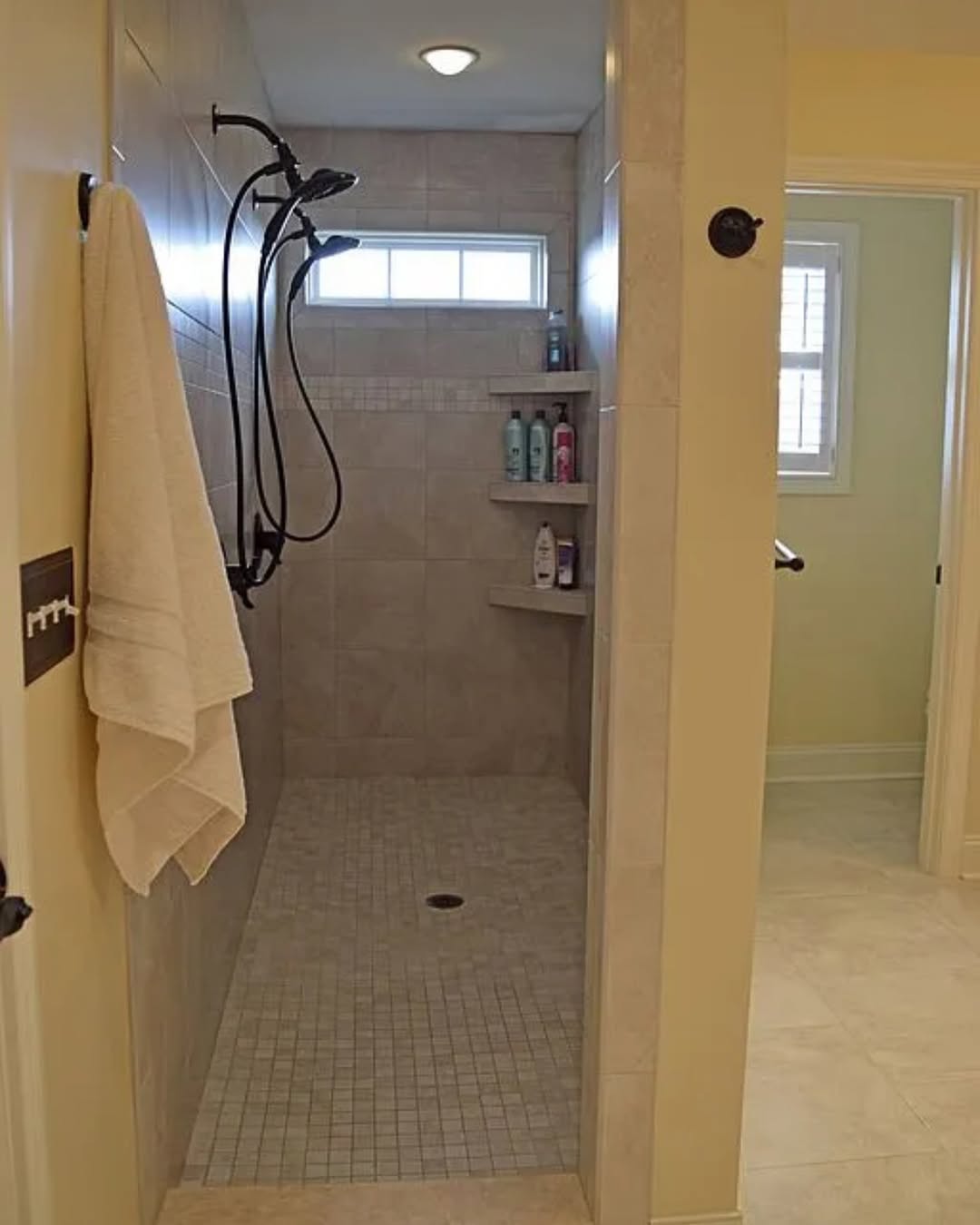 Walk-in shower with beige tile walls and floor, two built-in corner shelves for toiletries, a towel hanging on a nearby hook, and a small horizontal window above—perfect no door shower inspiration for small bathrooms.