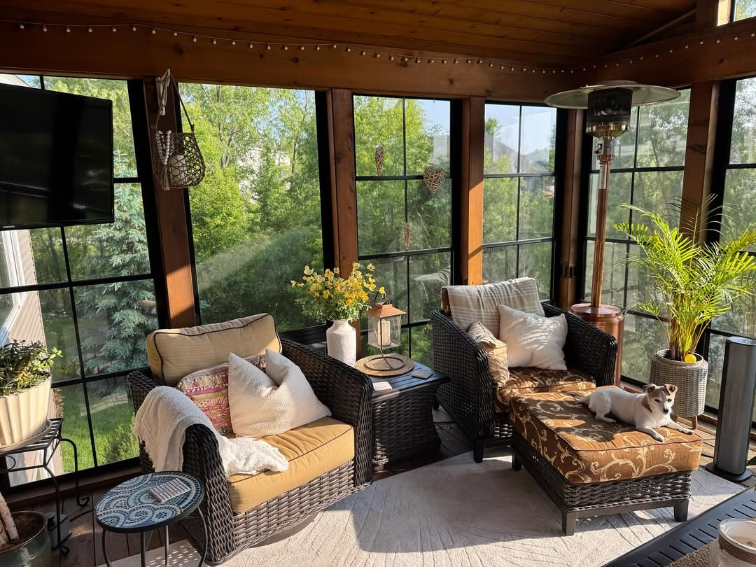 A sunroom with wicker chairs, patterned cushions, a small table with a vase, potted plants, and a dog lying on an ottoman—perfect for gathering 3 season porch ideas. Large windows overlook lush greenery outside.