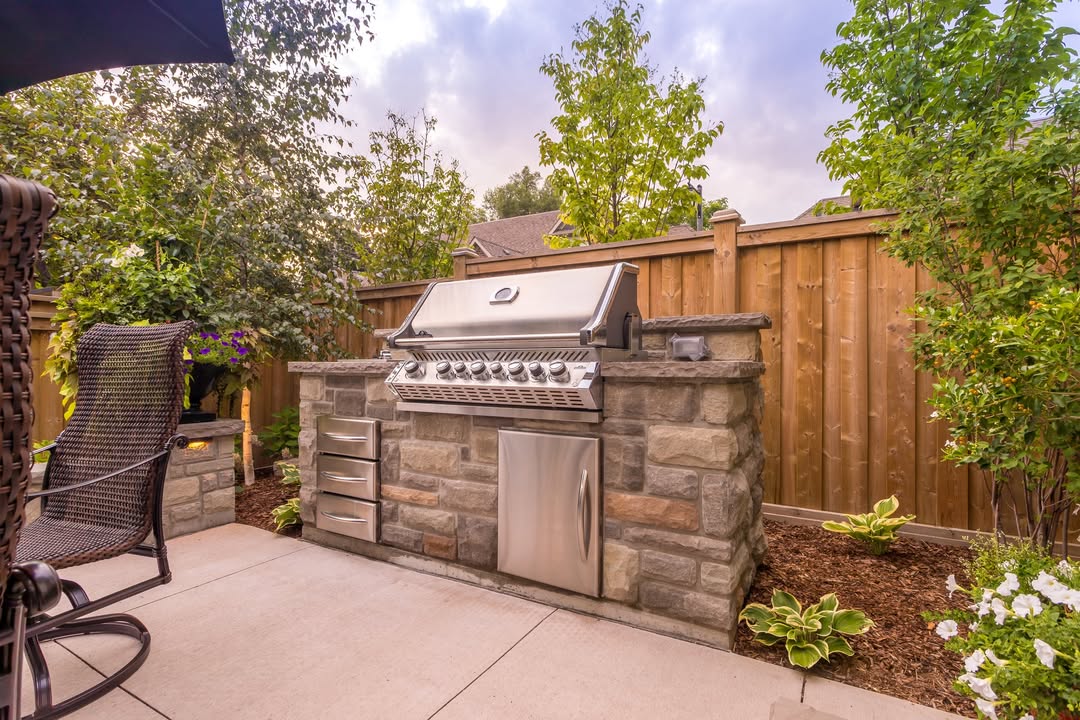 This rustic outdoor kitchen features a built-in stainless steel gas grill with storage drawers and a mini fridge, surrounded by stone on a patio with plants and a wooden fence in the background. Perfect for inspiring outdoor kitchen ideas.
