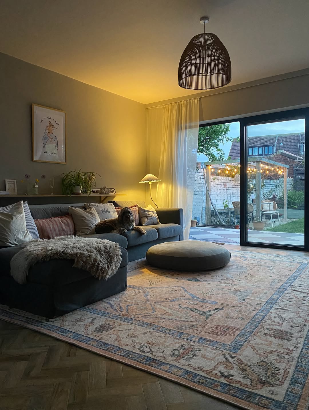A cozy living room with a gray sofa, a dog, a round ottoman, living room decor accents, a patterned rug, and sliding glass doors opening to a garden with string lights.