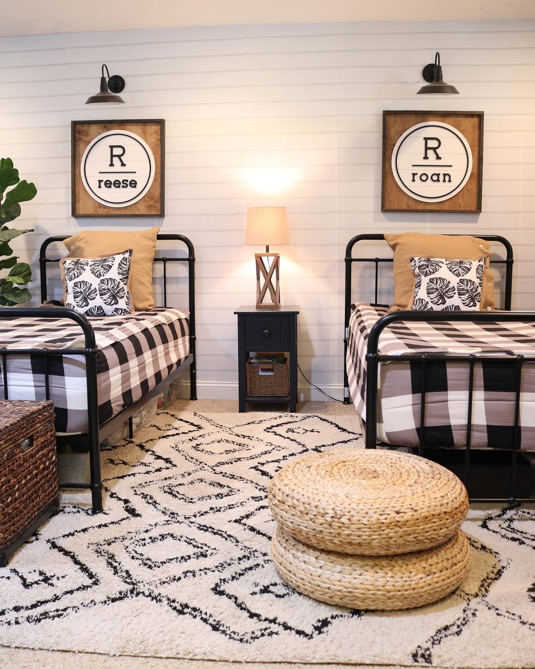 Two twin beds with black frames, buffalo plaid bedding, and personalized name art above each bed, separated by a nightstand with a lamp, create a cozy kids room design in this well-lit bedroom.