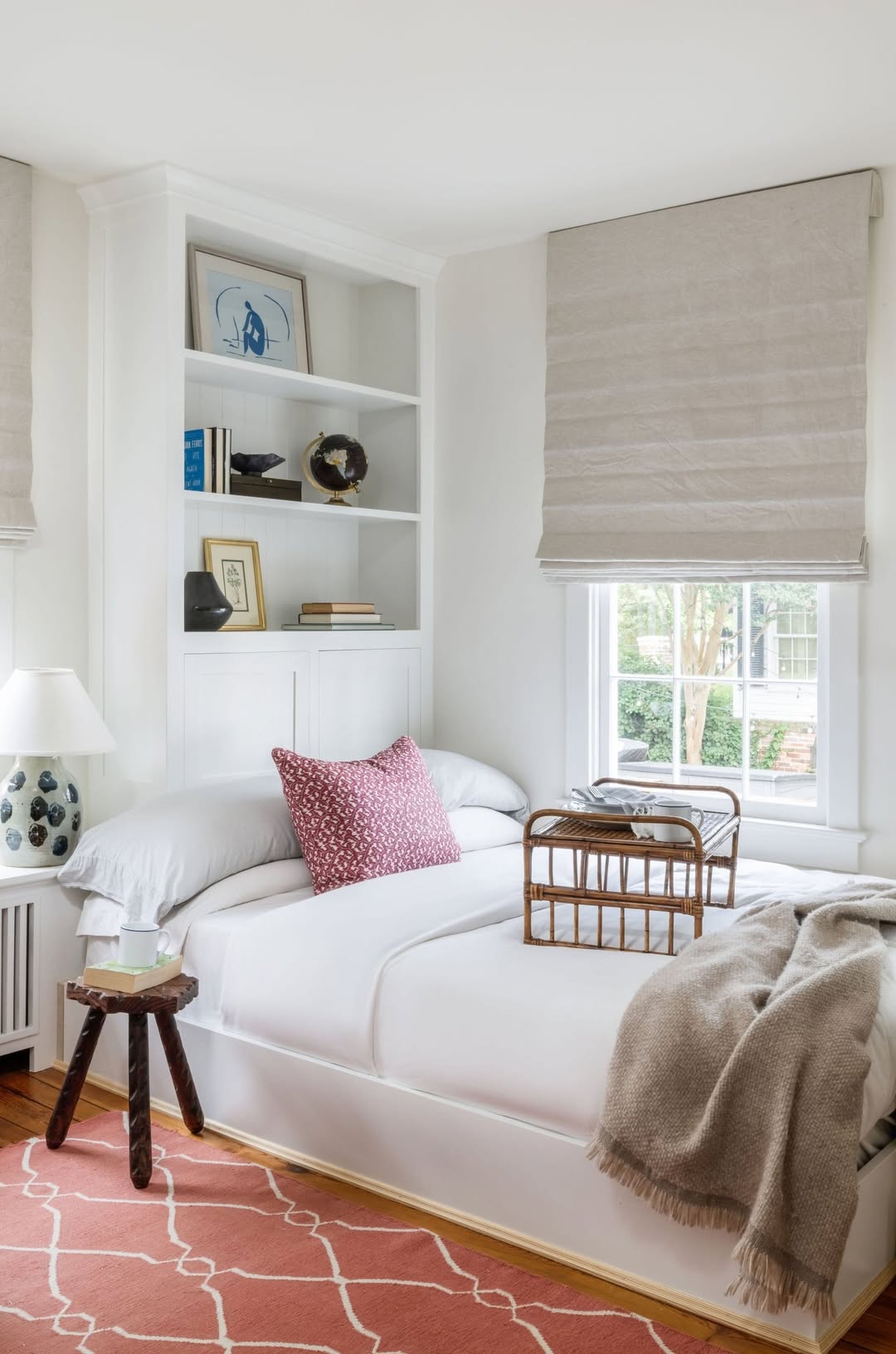 A neatly made bed with white bedding, a pink patterned pillow, and a beige throw blanket sits beside a window with Roman shades and built-in shelves—a perfect example of small apartment decor. A tray and lamp are on the side table.