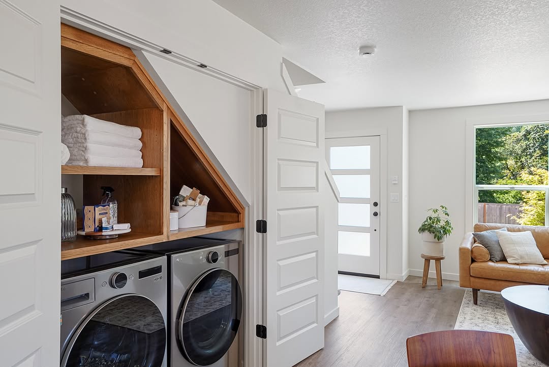 A modern under stair laundry room with a washer, dryer, shelves with towels, and cleaning supplies is cleverly built into a bright living space.