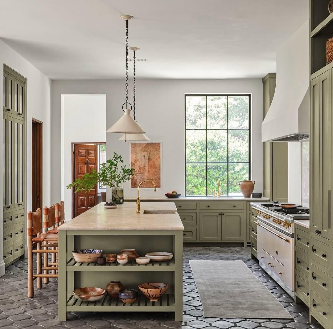 A spacious kitchen design with sage green cabinets, a large island, wooden stools, pendant lights, and a window overlooking greenery. Patterned floor tiles and open shelves displaying ceramics inspire modern open kitchen ideas.