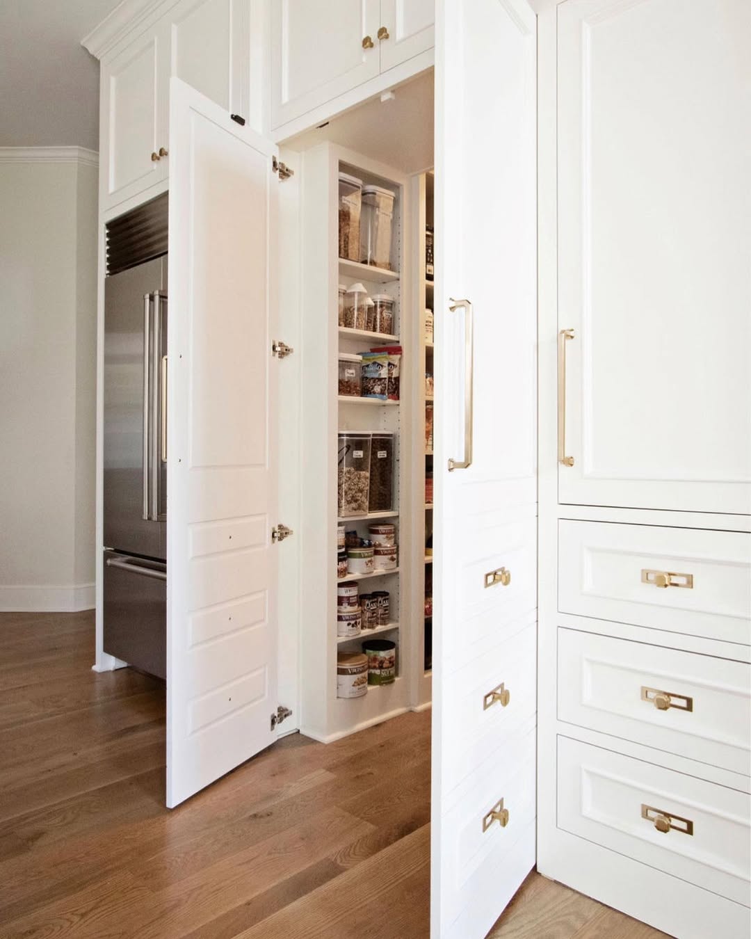 A built-in white kitchen pantry with open double doors reveals organized shelves of food items next to a stainless steel refrigerator, delivering serious wow factor and inspiring real life pantry door ideas.