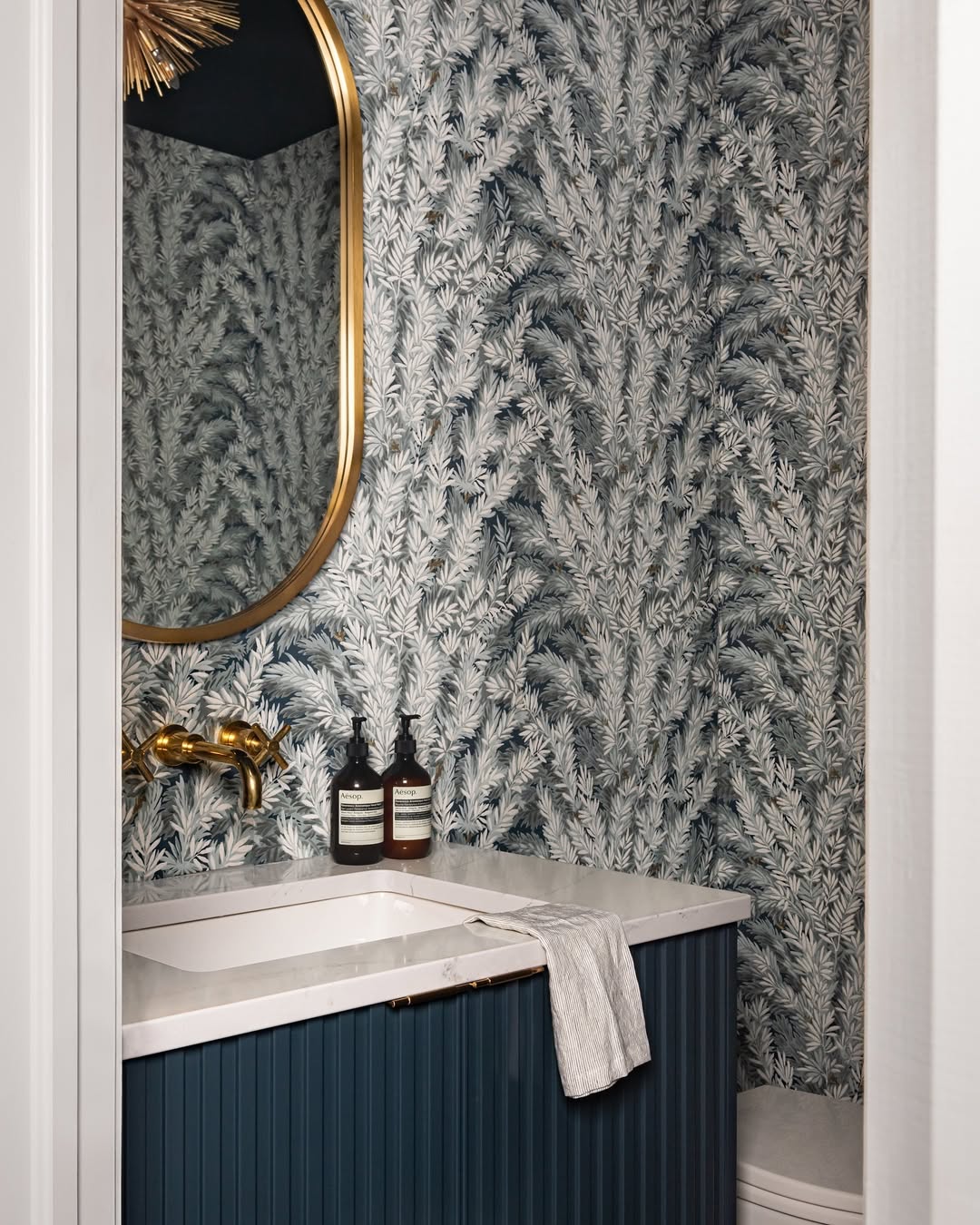 A bathroom with leafy wallpaper, a gold-framed oval mirror, gold faucet, blue vanity, white countertop, and a hand towel draped over the sink—perfect inspiration for stylish powder rooms with serious wow-factor.