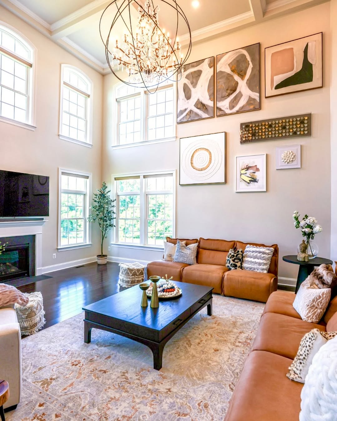 A bright, modern high ceiling living room with tall windows, tan leather sofas, a black coffee table, wall art, a chandelier, and a fireplace with a TV above it—an inspiring space to make your home bigger.