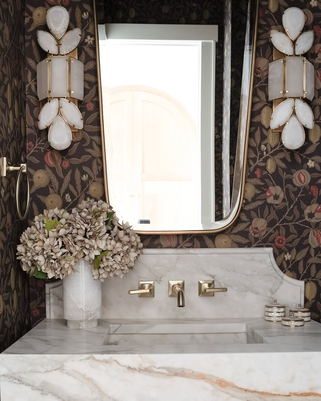 A marble bathroom vanity with gold fixtures, a vase of dried flowers, decorative wall sconces, and floral wallpaper—perfect inspiration for stylish powder rooms that deliver instant wow-factor.