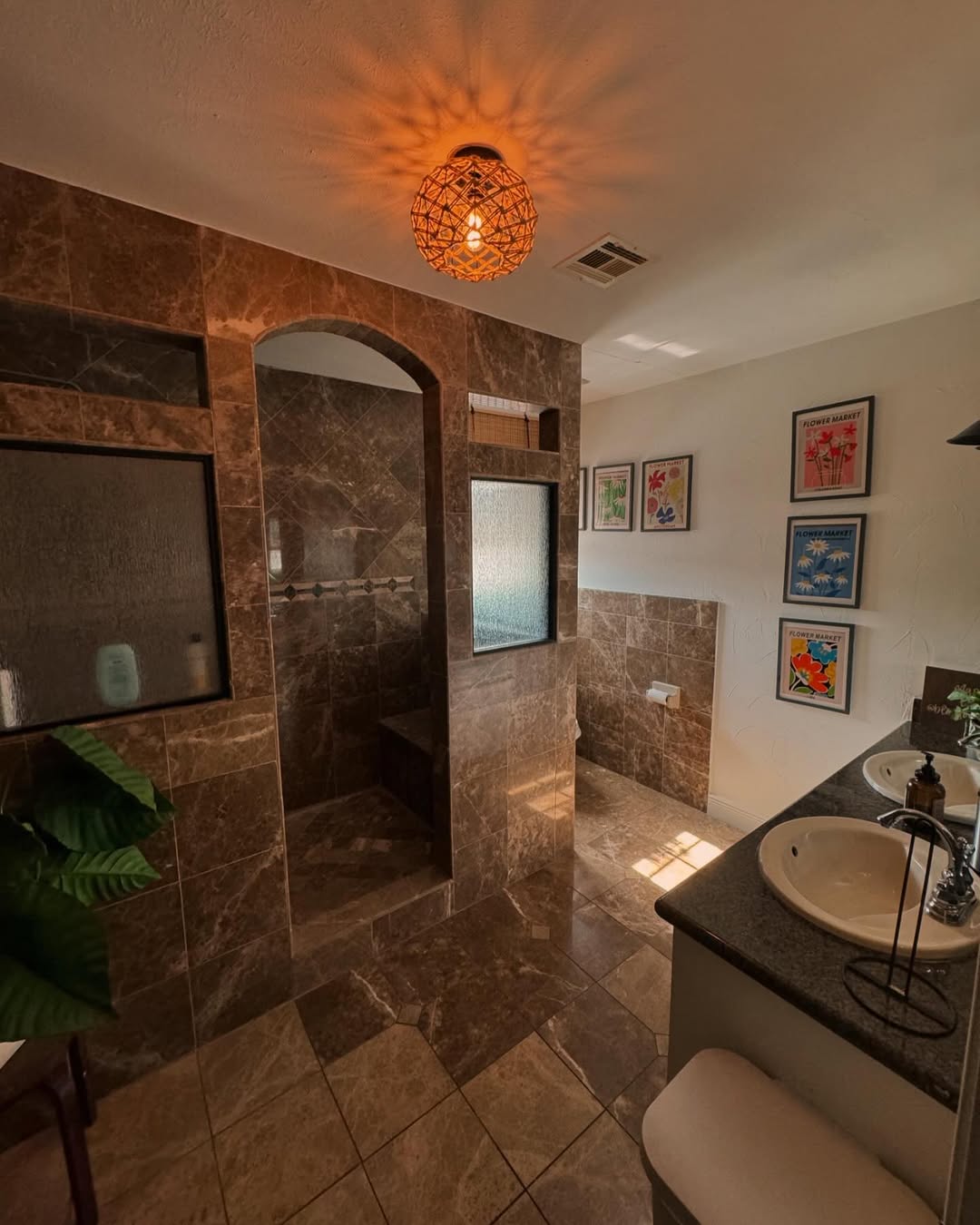 A bathroom with brown marble tiles, a no door walk-in shower—ideal for small bathrooms—double sinks, framed wall art, a decorative ceiling light, and a plant in the corner.
