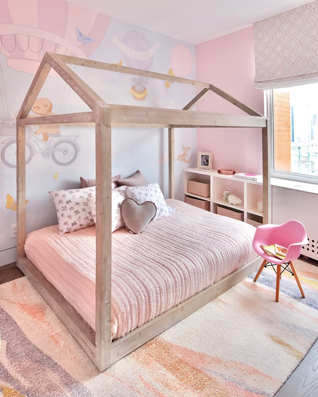 This cute toddler bedroom features a house-shaped wooden bed frame with pink bedding and pillows. Dreamy wall art showcases vintage vehicles. A pink chair and shelving cozily sit in the corner, all bathed in natural light from the large window on the right.