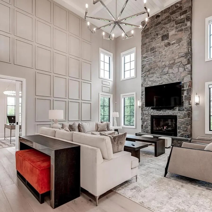Spacious high ceiling living room with a stone fireplace, mounted TV, large windows, neutral sofas, modern chandelier, and a red bench under a console table—perfect for those seeking inspiring high ceiling ideas.