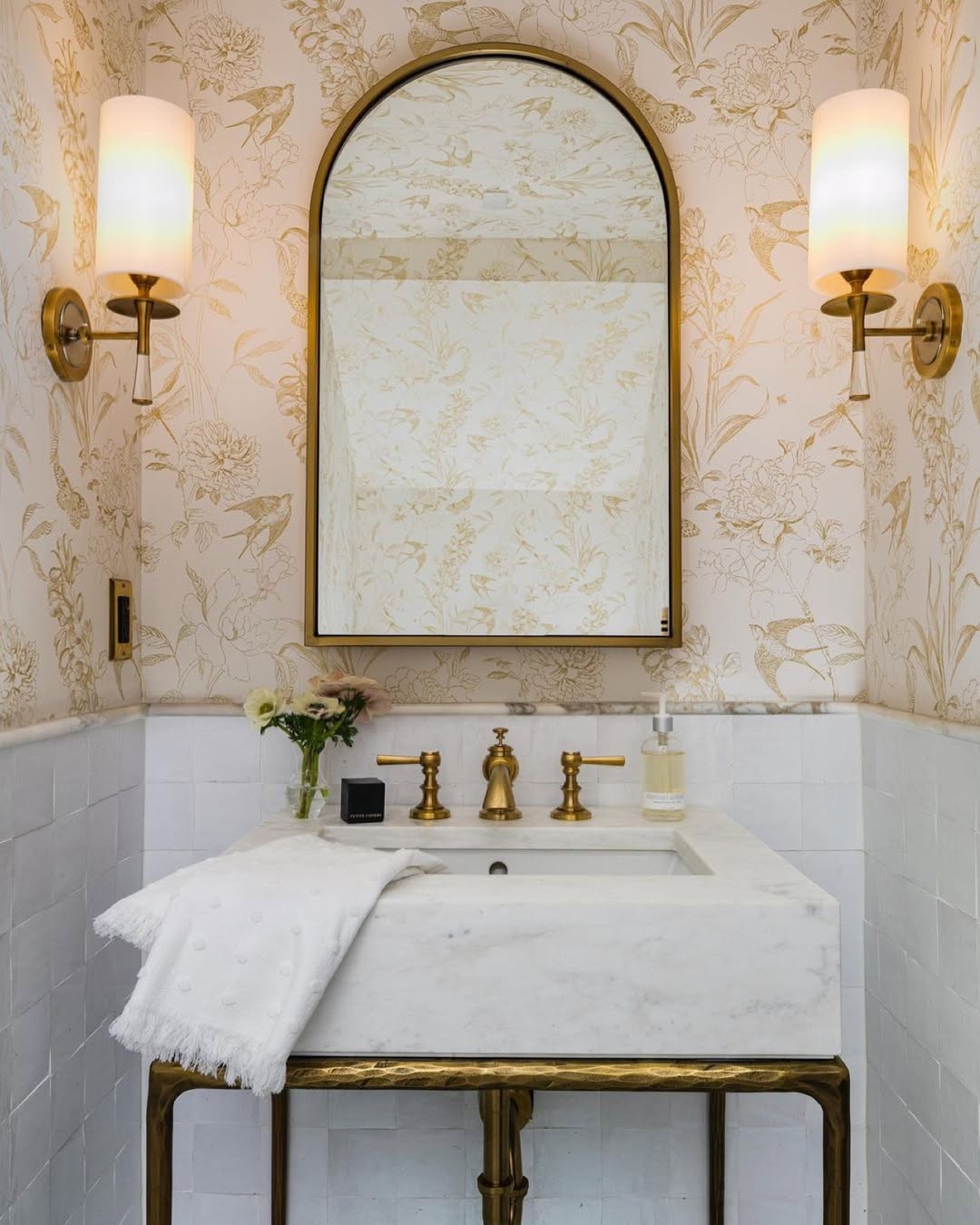 A small bathroom vanity with a marble sink, gold fixtures, wall mirror, two sconces, floral wallpaper, towel, flowers, and soap dispenser—perfect inspiration for anyone seeking wow-factor powder room ideas.
