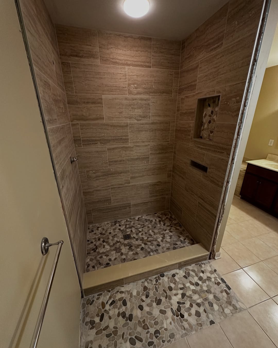 Walk-in shower with tan tile walls, pebble-style floor, built-in shelf, and overhead lighting. This no door walk-in shower is perfect for small bathroom ideas, with an adjacent beige tile floor and partial view of the sink cabinet.