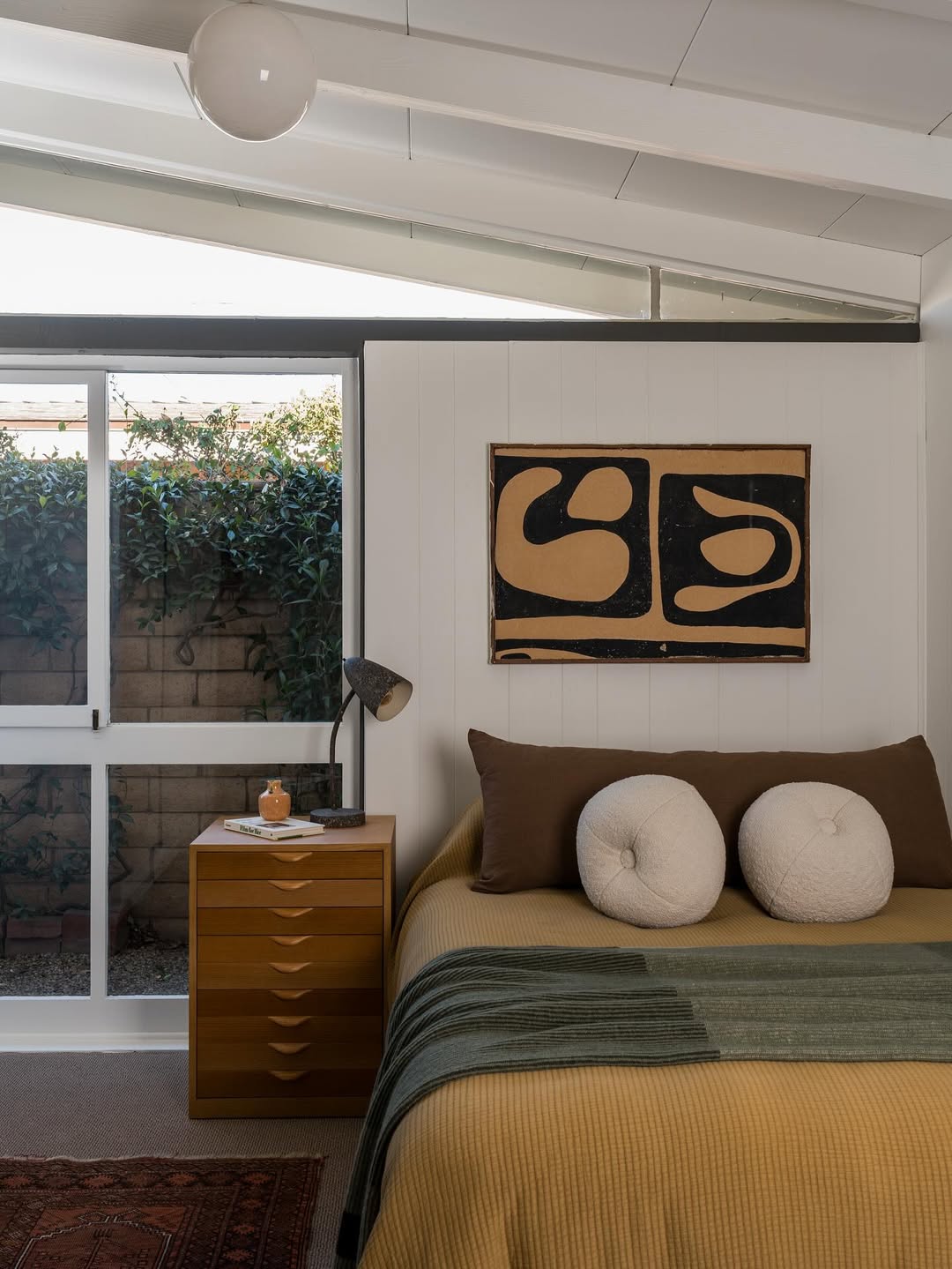 A midcentury modern bedroom with a mustard bedspread, two round pillows, abstract wall art, and a wooden nightstand features Ranch House Decor touches and a glass door revealing lush greenery outside.