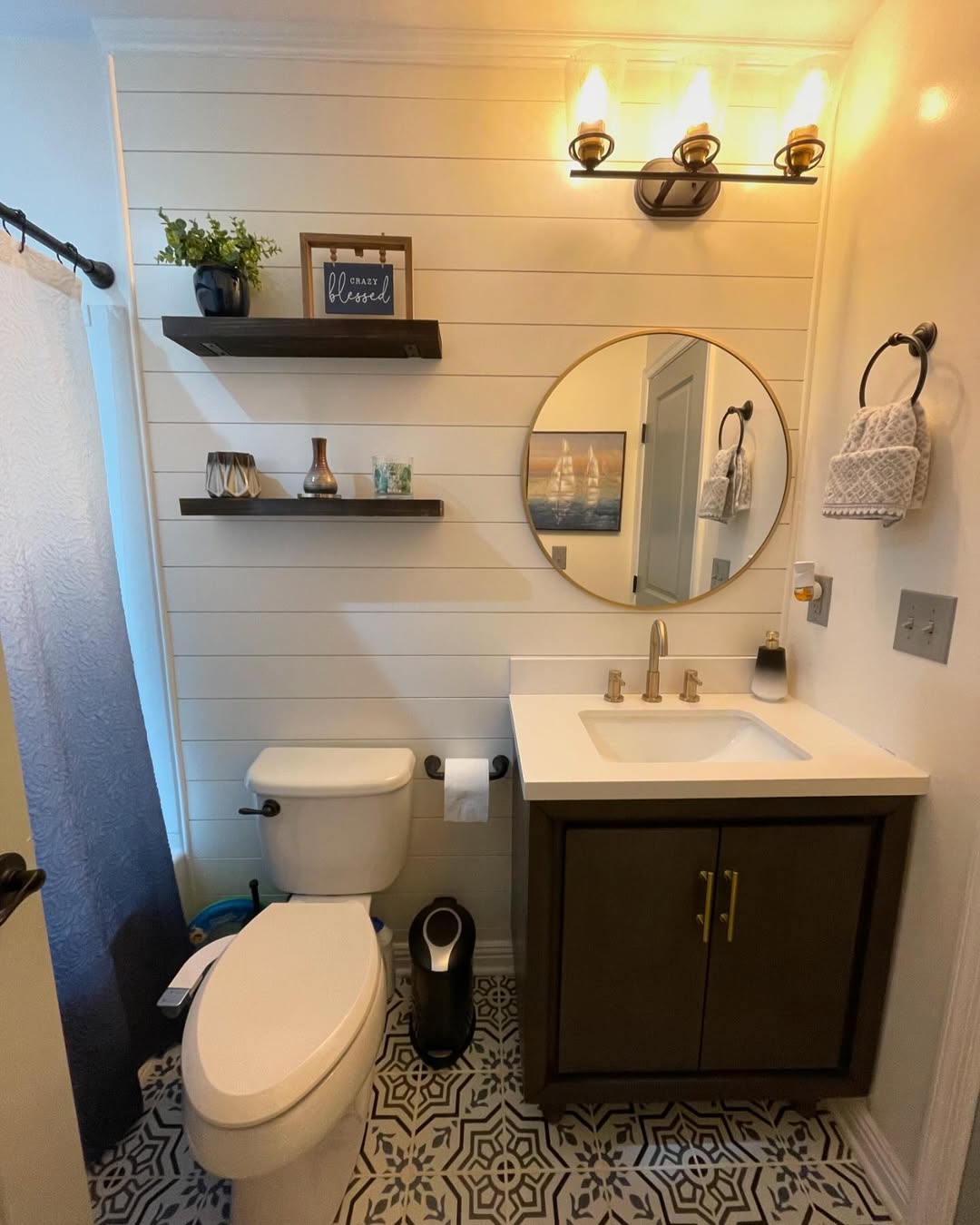 A modern shiplap wall bathroom features a round mirror, dark vanity, shelves with decor, white shiplap walls, toilet, patterned tile floor, and a wall-mounted light fixture.