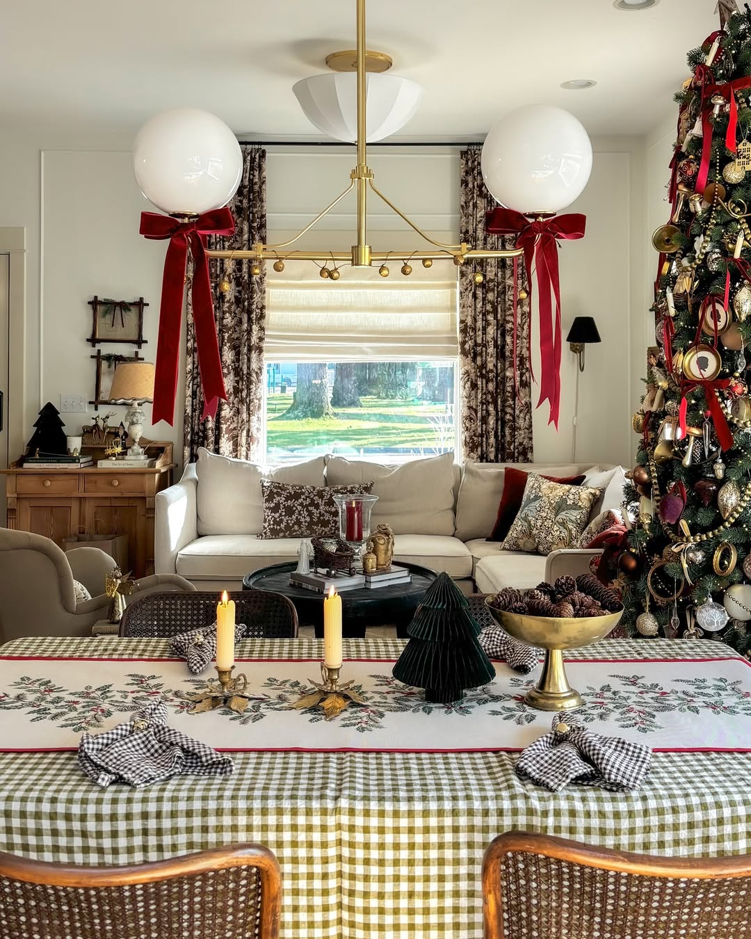A living room decorated for Christmas with a large tree, a set dining table with candles, festive tablecloth, and elegant Christmas chandelier decor above, plus a sofa in the background near large windows with curtains.