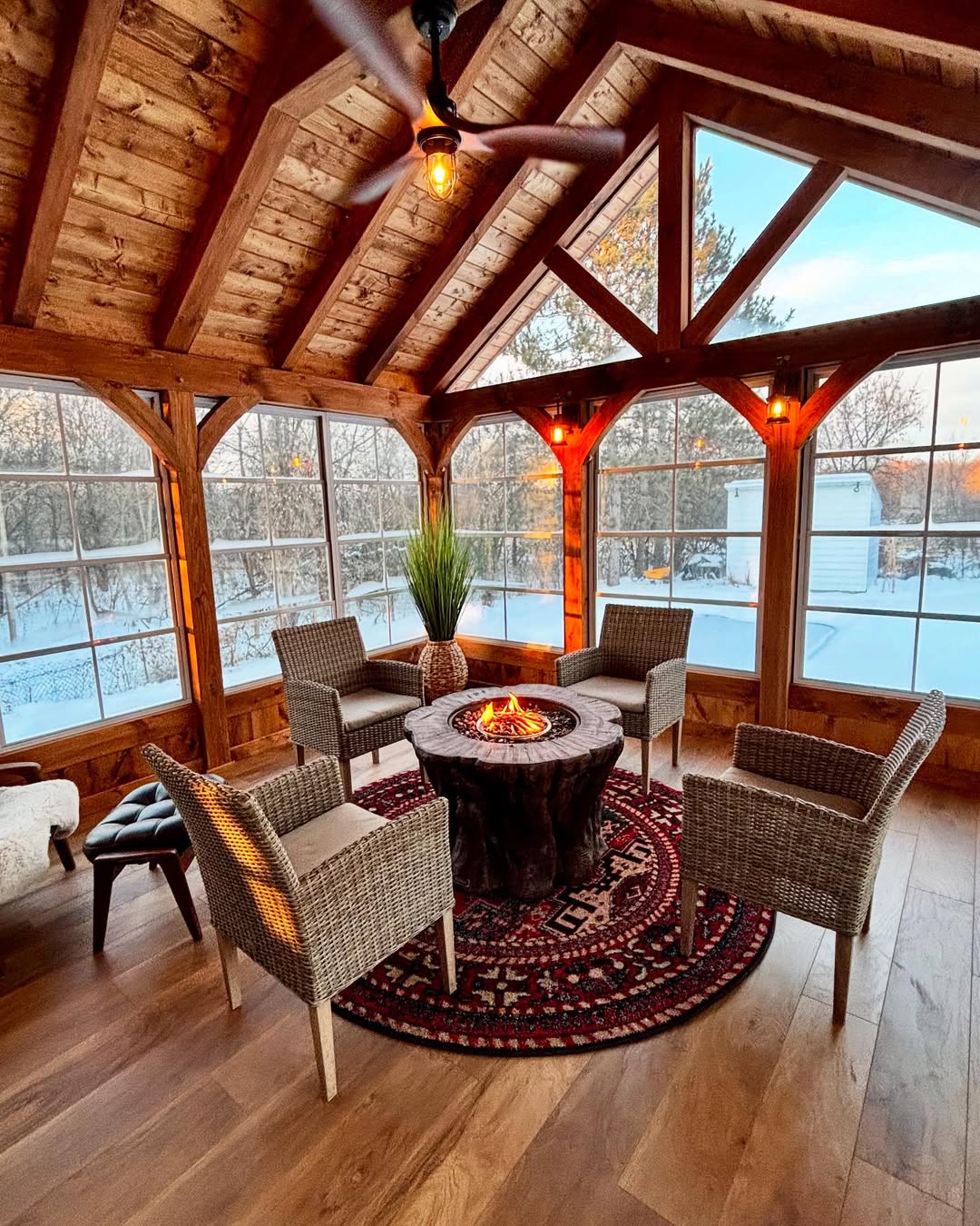 A cozy seasonal porch with wood beams features four wicker chairs around a circular fire pit on a patterned rug, with large windows showing a snowy landscape—perfect inspiration for 3 season porch ideas.