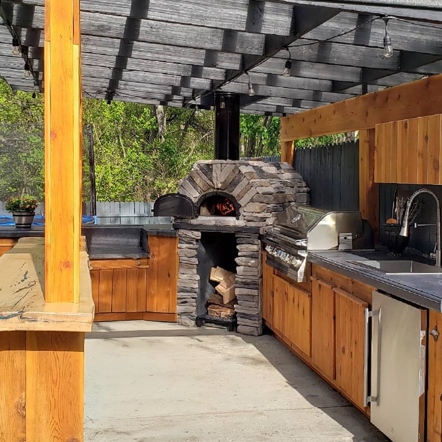 This Rustic outdoor kitchen features a wood-fired stone oven, grill, stainless steel sink, and wooden cabinetry under a pergola. Logs are neatly stored below the oven, making it perfect for those seeking rustic kitchen ideas amid nature.