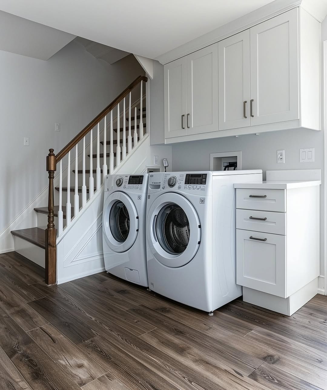 A modern under stair laundry room features a front-loading washer and dryer beneath sleek white cabinets, all set beside a staircase with wood flooring throughout the space.