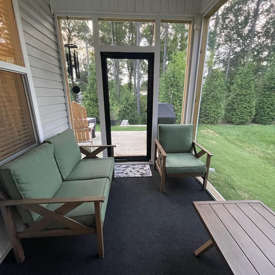 A screened porch with green cushioned seating, a wooden coffee table, and a view of a backyard with grass, trees, and a patio area through a glass door—perfect for those seeking inspiring 3 season porch ideas.