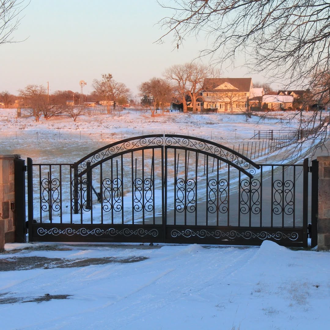 A wrought iron farmhouse gate stands over a snow-covered driveway, with a farmhouse and trees visible in the background.