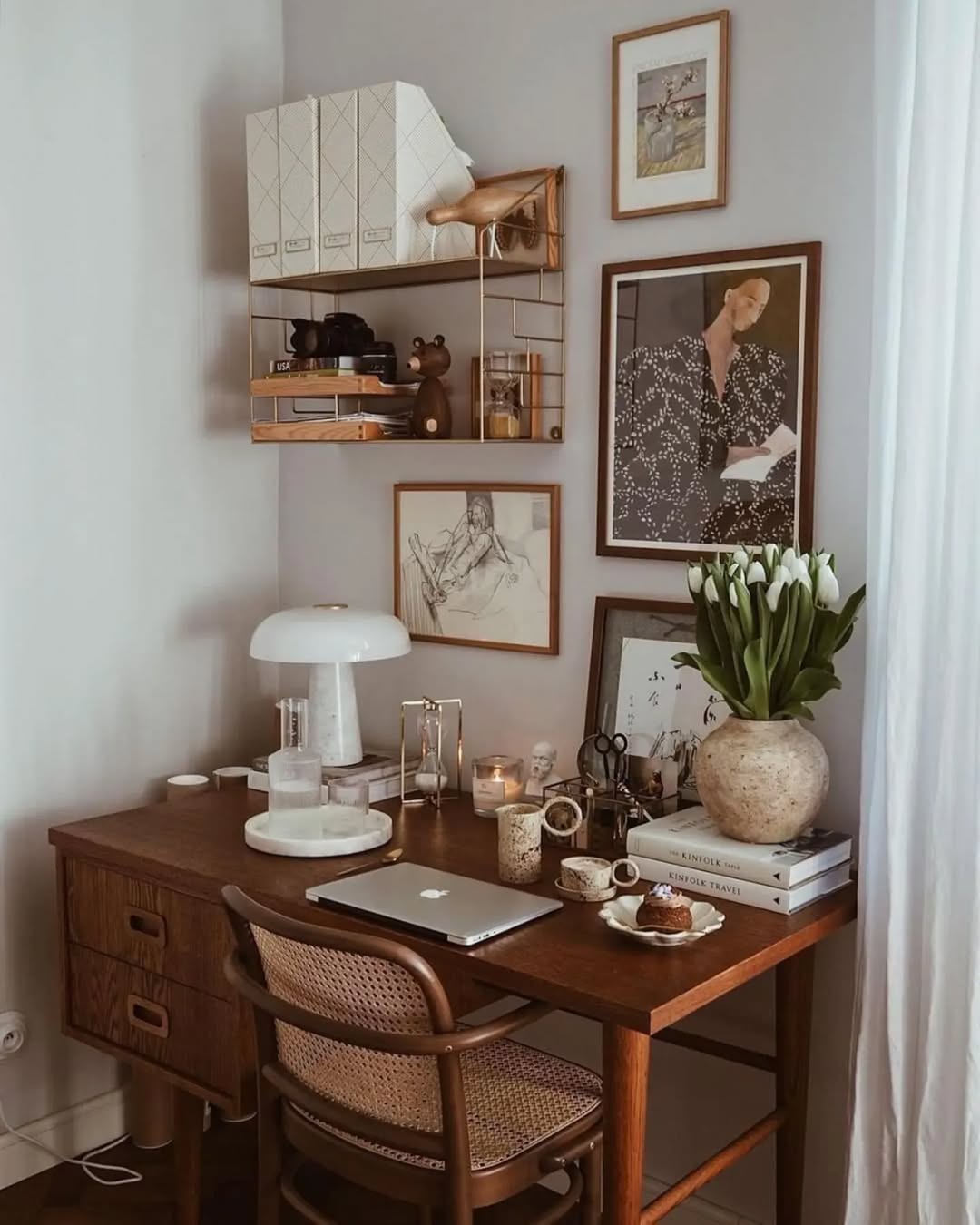 A wooden desk with a laptop, lamp, vase of white tulips, books, and boho decor creates a cozy Boho home office vibe. Set against a wall with shelves and framed artwork, a wooden chair completes this stylish home office idea.