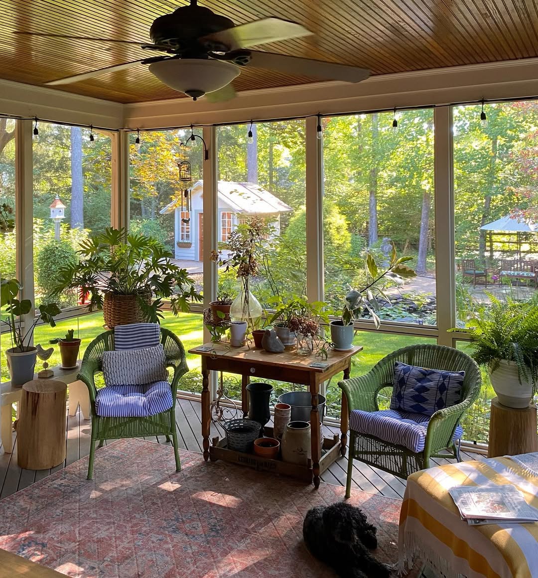 A sunlit seasonal porch with wicker chairs, potted plants, a small table, and a ceiling fan overlooks a green garden with trees and a white garden shed—perfect for cozy porch decor.