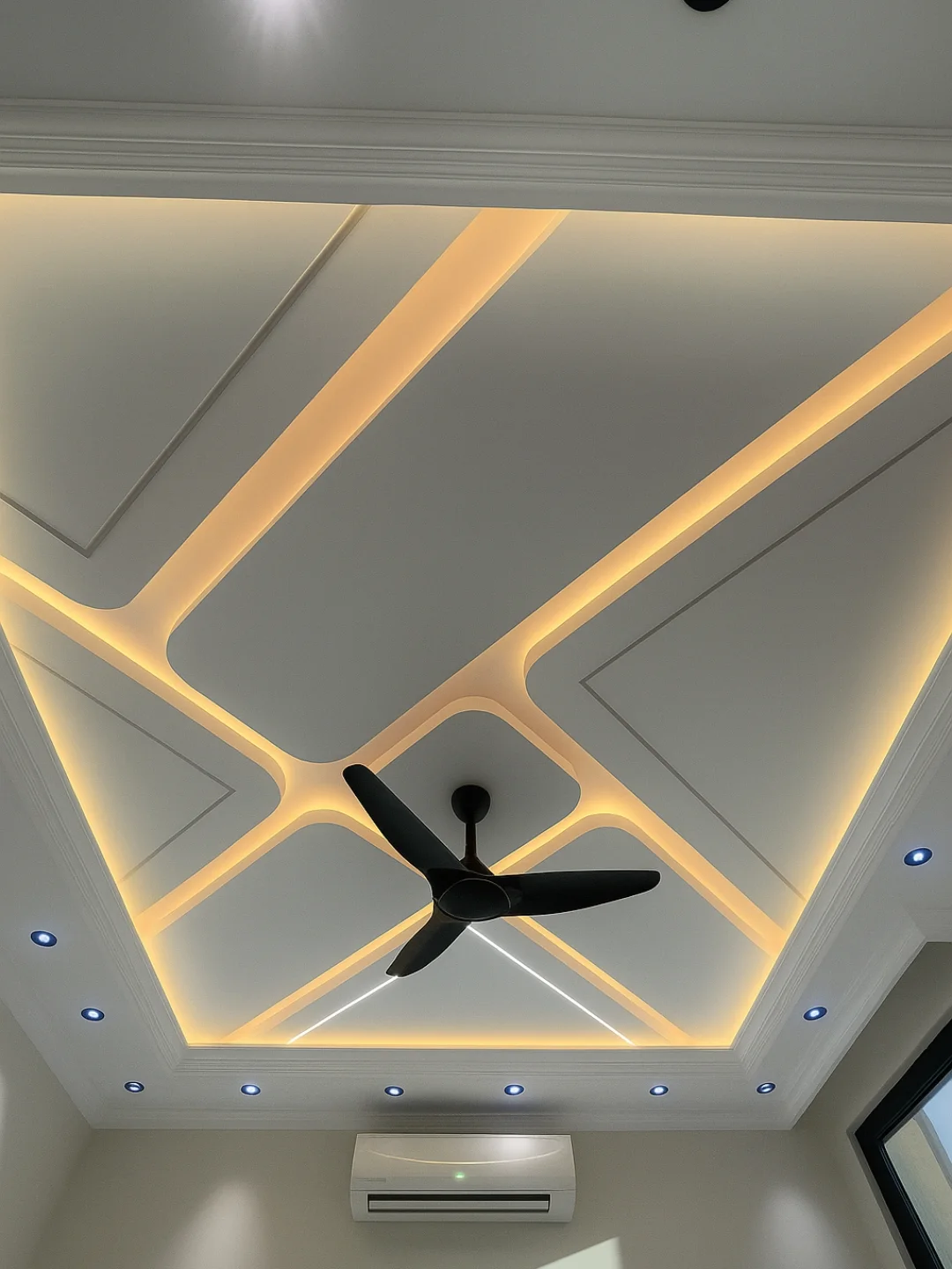 A modern ceiling with geometric recessed lighting, a black ceiling fan in the center for sleek Home Decor, blue spotlights around the perimeter, and an air conditioner mounted on the wall below. Perfect for those seeking unique Ceiling Fan Ideas.
