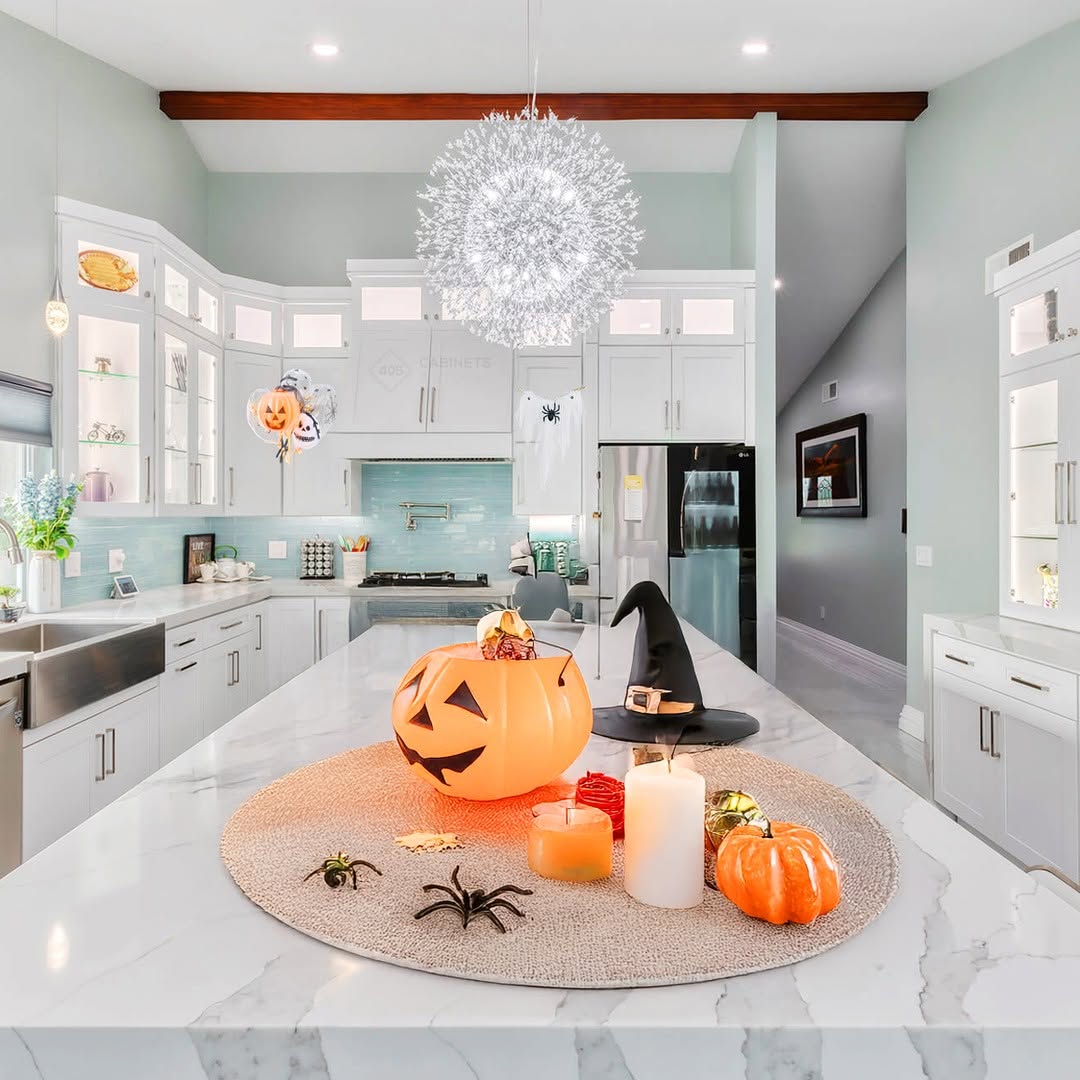 Modern kitchen with white cabinets, a marble island, and creative Halloween kitchen decorations like pumpkins, candles, a witch hat, and fake spiders arranged on the countertop for festive flair.