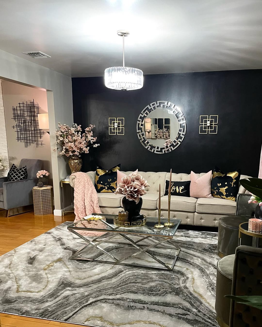 Modern black living room featuring a beige sofa, black accent wall, decorative pillows, glass coffee table, floral arrangements, and a round mirror above the sofa.