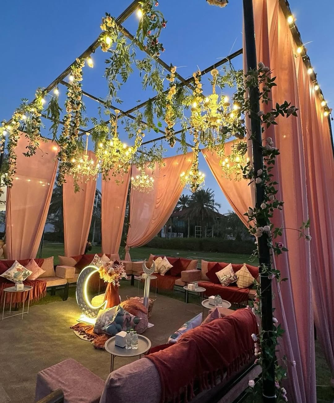 Chic patio decoration featuring pink drapes, string lights, hanging greenery, and floral arrangements, complemented by cushioned seating and a glowing letter