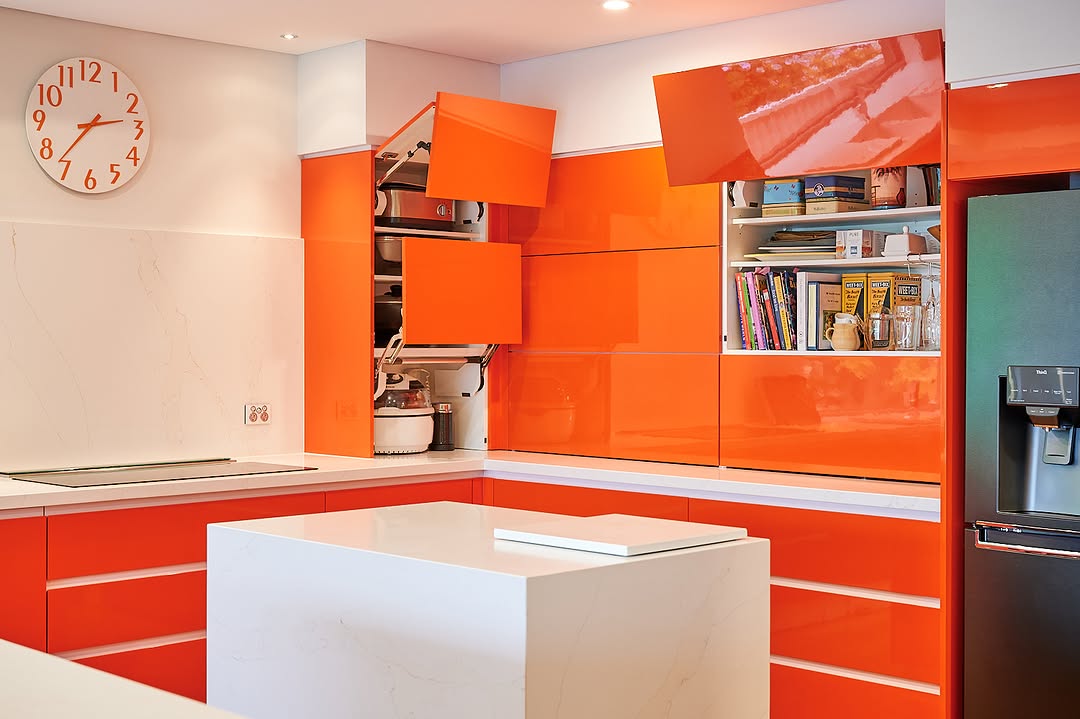 Modern orange kitchen with glossy cabinets—some open to reveal appliances and shelves—a white countertop island, and a wall clock showing 2:19.