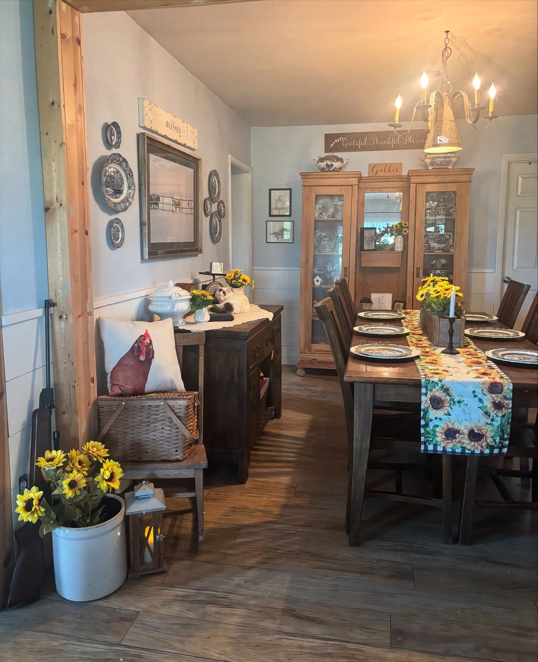 A farmhouse-style dining room with a wooden table set for six, sunflower accents, a sideboard, a china cabinet, wall art, and touches of vintage farmhouse decor for a cozy, inviting atmosphere.