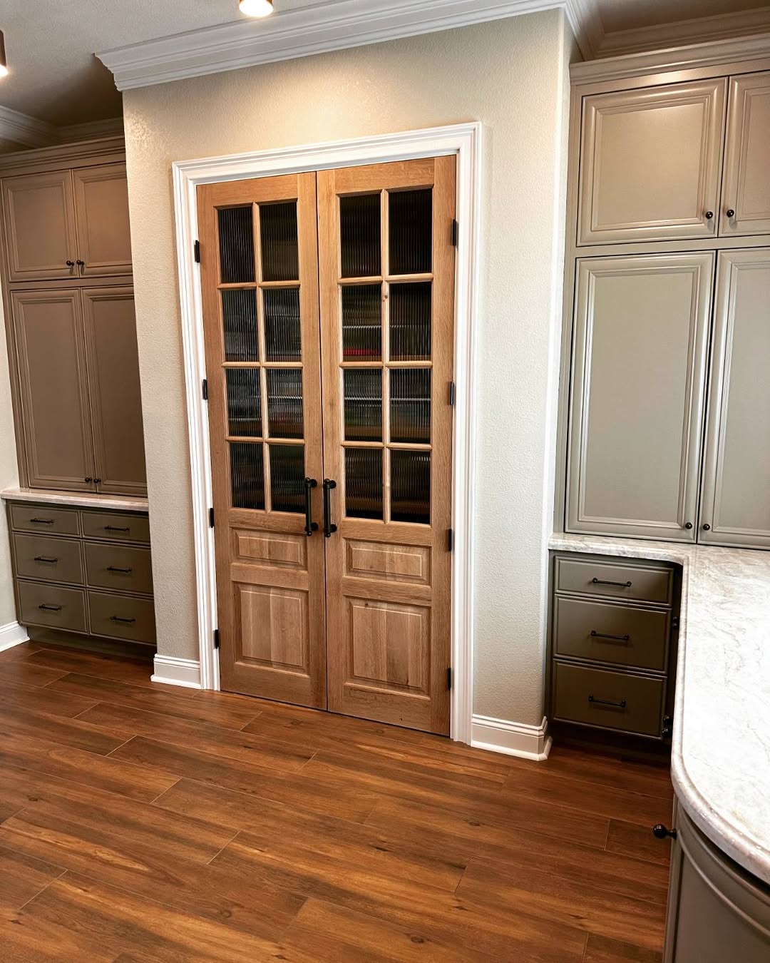 A real life kitchen features wood flooring, taupe cabinets, and double wooden pantry doors with glass panels and black handles—delivering serious wow factor and great pantry door ideas.