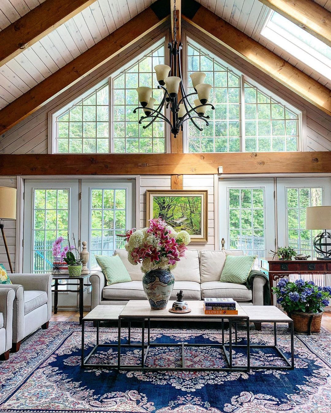 A spacious living room with vaulted ceilings, large windows, a white sofa, a floral rug, and a coffee table with a large vase of flowers—perfect high ceiling decor to make your home feel bigger and brighter.
